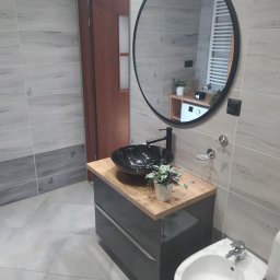 Maryka Ryszard Kapałka - Nowoczesna łazienka z szarą zabudową umywalkową, czarną misą umywalki na drewnianym blacie i okrągłym lustrem z czarną ramą, widoczny bidet i płytki imitujące drewno.