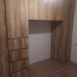 Maryka Ryszard Kapałka - Drewniana szafa wnękowa z szufladami i pustą wnęką na biurko lub toaletkę, oświetlona od góry.