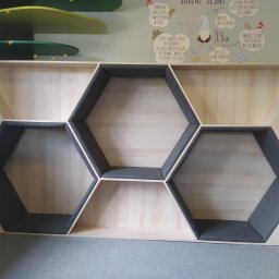 Maryka Ryszard Kapałka - Niska, drewniana szafka z sześciokątnymi wnękami wyłożonymi szarą tapicerką, umieszczona w pokoju dziecięcym z dekoracjami na ścianie.