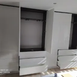 Biała, lakierowana szafa z otwartymi drzwiami i szufladami, widoczne drążki na ubrania i mechanizmy szuflad, w tle rolety okienne.