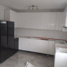 Maryka Ryszard Kapałka - Wnętrze kuchni w trakcie montażu, widoczne białe szafki bez blatów, czarna lodówka side-by-side i fragment marmurowej podłogi.