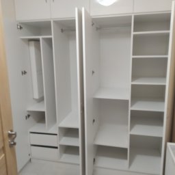 Maryka Ryszard Kapałka - Biała szafa wnękowa z otwartymi drzwiami, widoczne półki, drążki na ubrania i dwie szuflady w dolnej części.