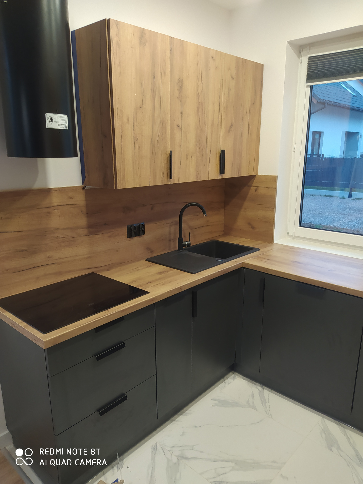 Nowoczesna kuchnia z ciemnymi szafkami dolnymi i górnymi szafkami z drewnopodobnym fornirem, czarnym zlewem i płytą grzewczą, oraz marmurową podłogą.