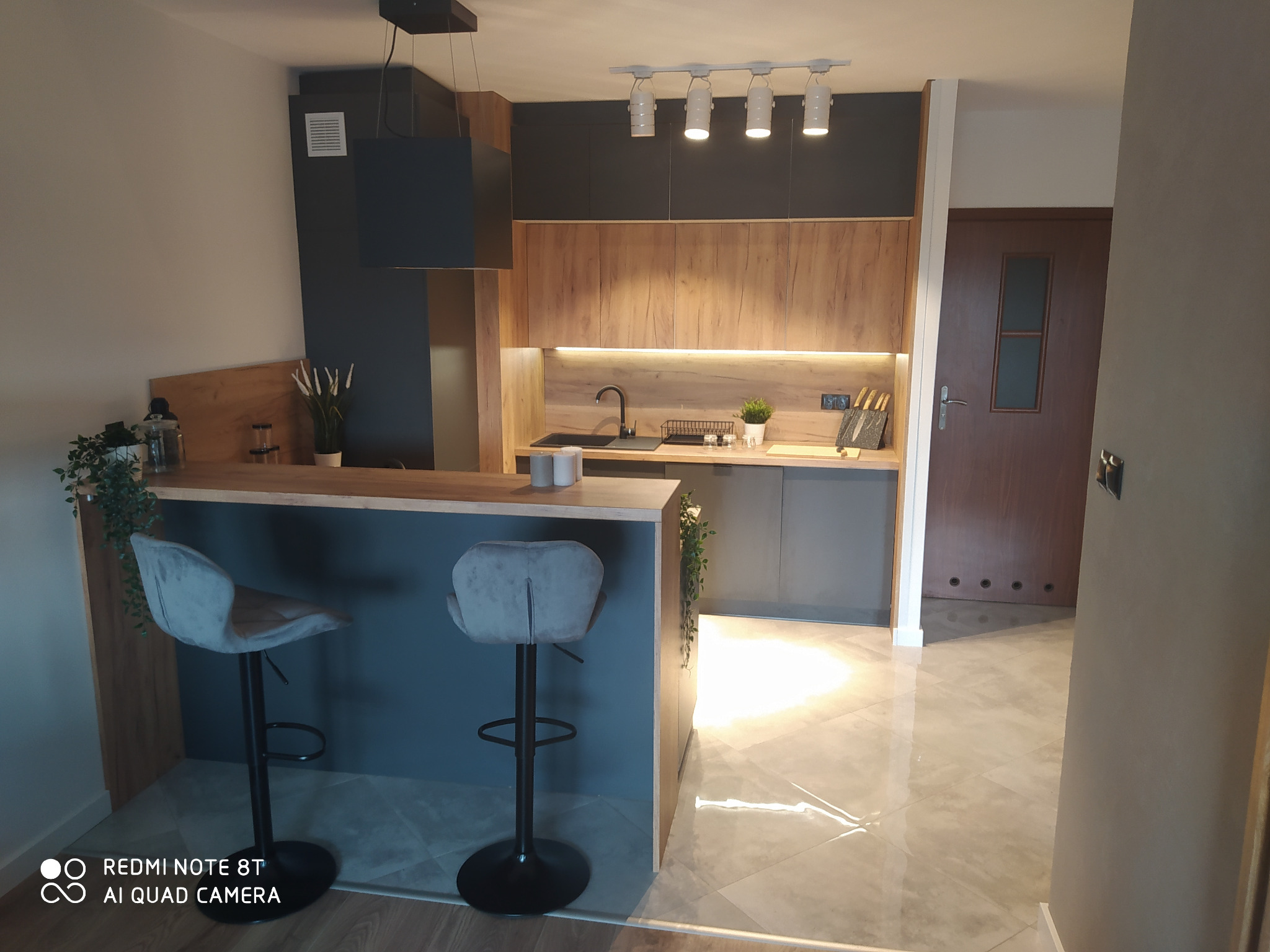 Nowoczesna kuchnia z drewnianymi frontami szafek górnych i ciemnoszarymi dolnymi, wyspą z dwoma szarymi hokerami i oświetleniem punktowym.