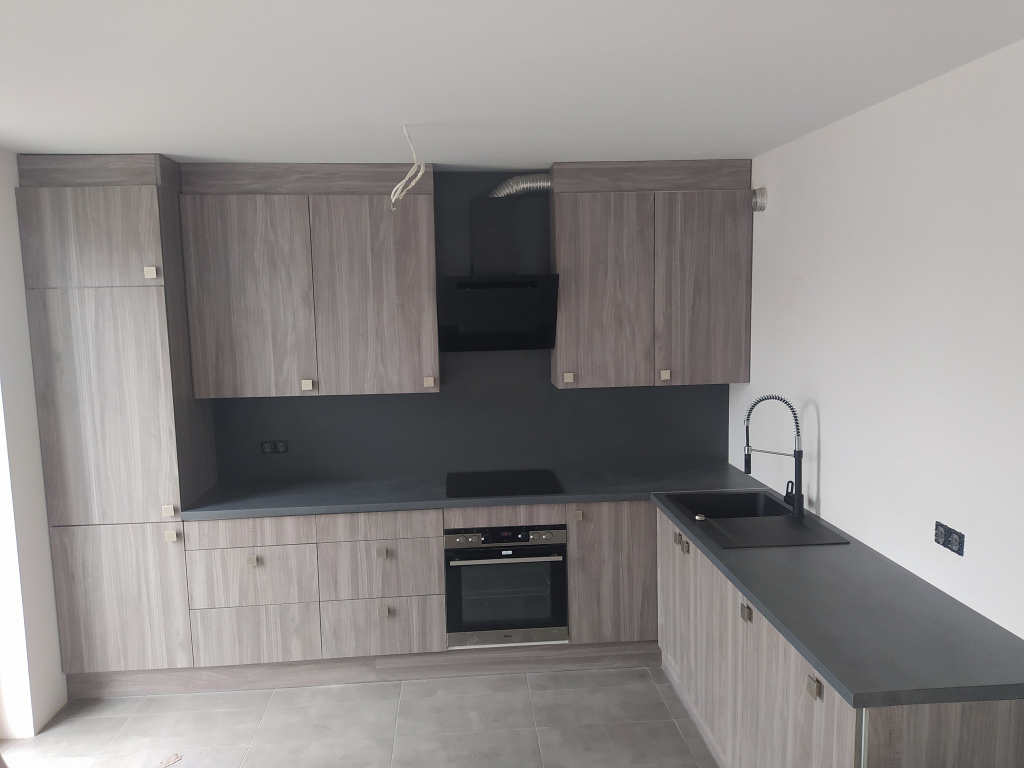 Nowoczesna kuchnia z szarymi frontami imitującymi drewno, czarnym blatem i sprzętem AGD w zabudowie, widoczne niedokończone podłączenie kabli