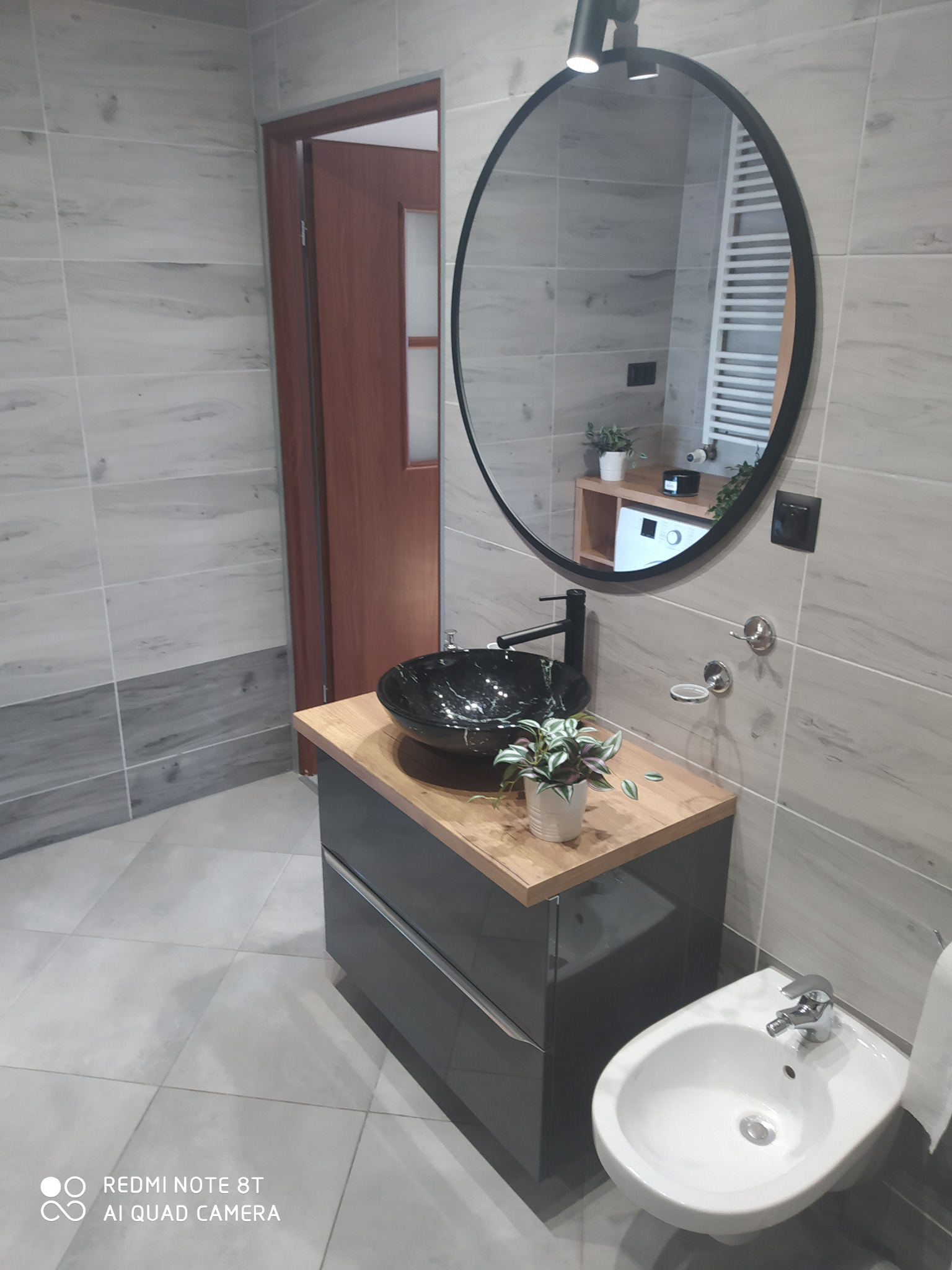 Nowoczesna łazienka z szarą zabudową umywalkową, czarną misą umywalki na drewnianym blacie i okrągłym lustrem z czarną ramą, widoczny bidet i płytki imitujące drewno.