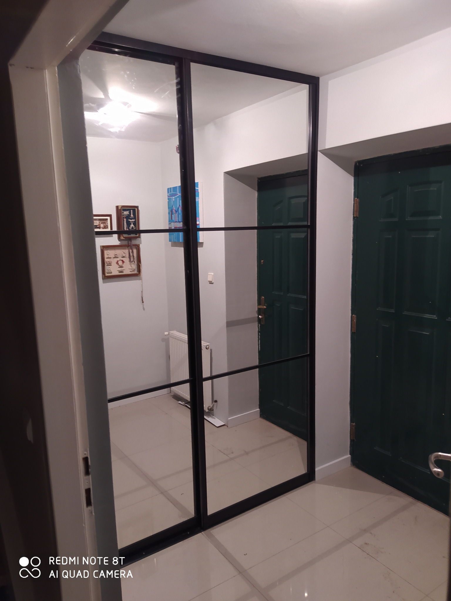 Przesuwne drzwi z czarną ramą i lustrzanym wypełnieniem, odbijające wnętrze przedpokoju z grzejnikiem i obrazami na ścianie.