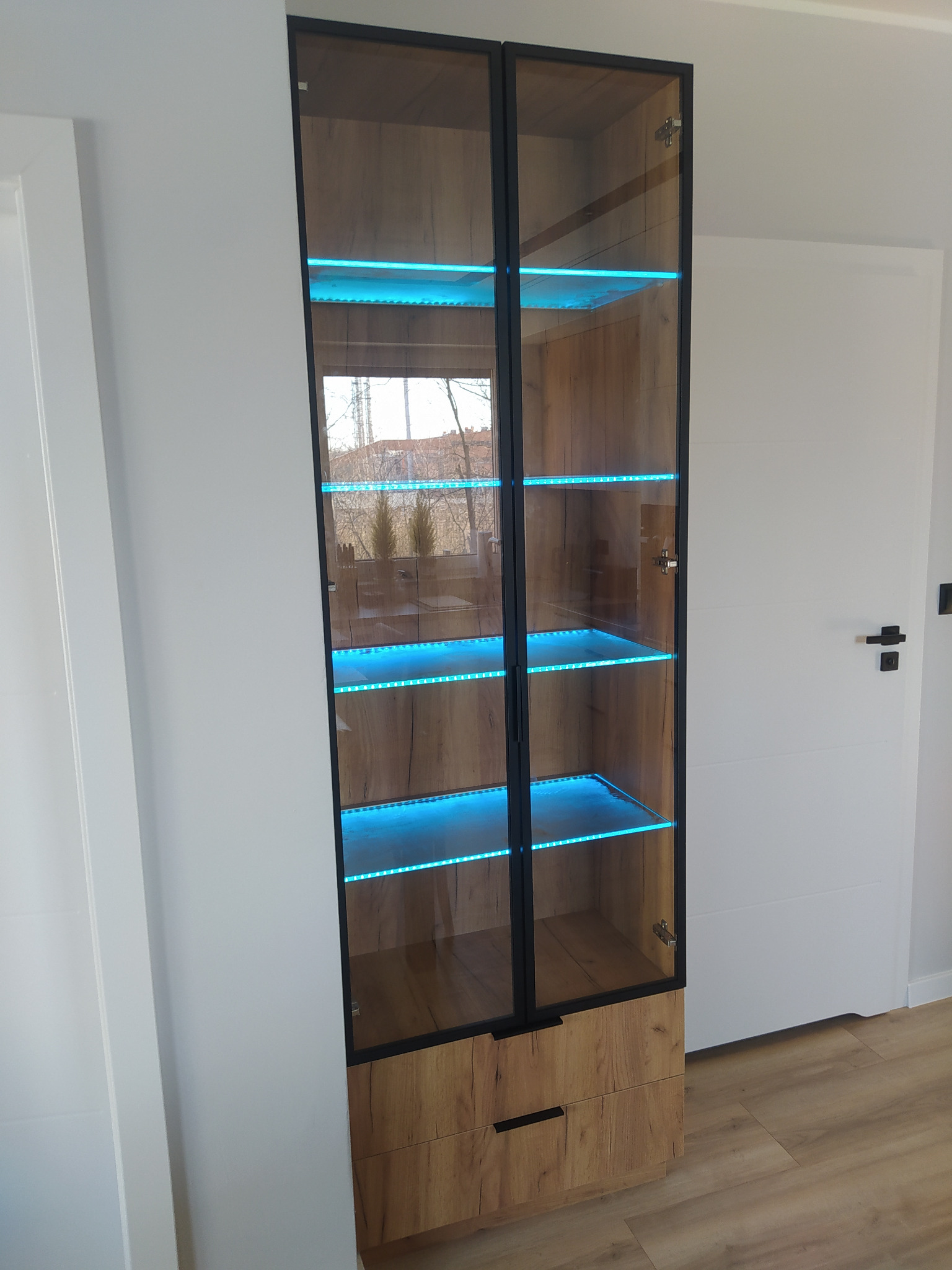 Wysoka, przeszklona witryna z drewnianym wykończeniem i szklanymi półkami podświetlonymi niebieskim światłem LED, ustawiona w rogu pokoju obok białych drzwi.