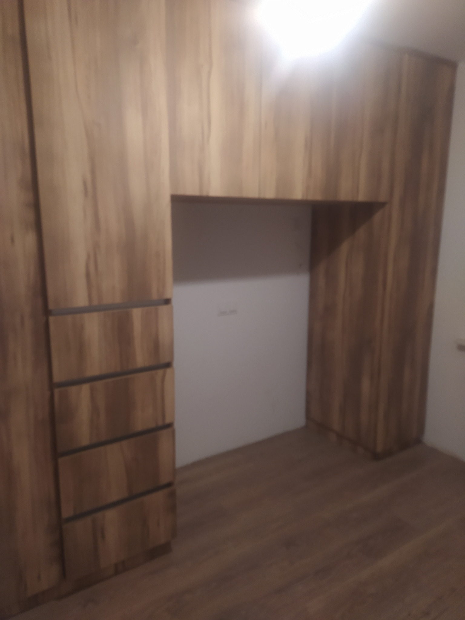 Drewniana szafa wnękowa z szufladami i pustą wnęką na biurko lub toaletkę, oświetlona od góry.