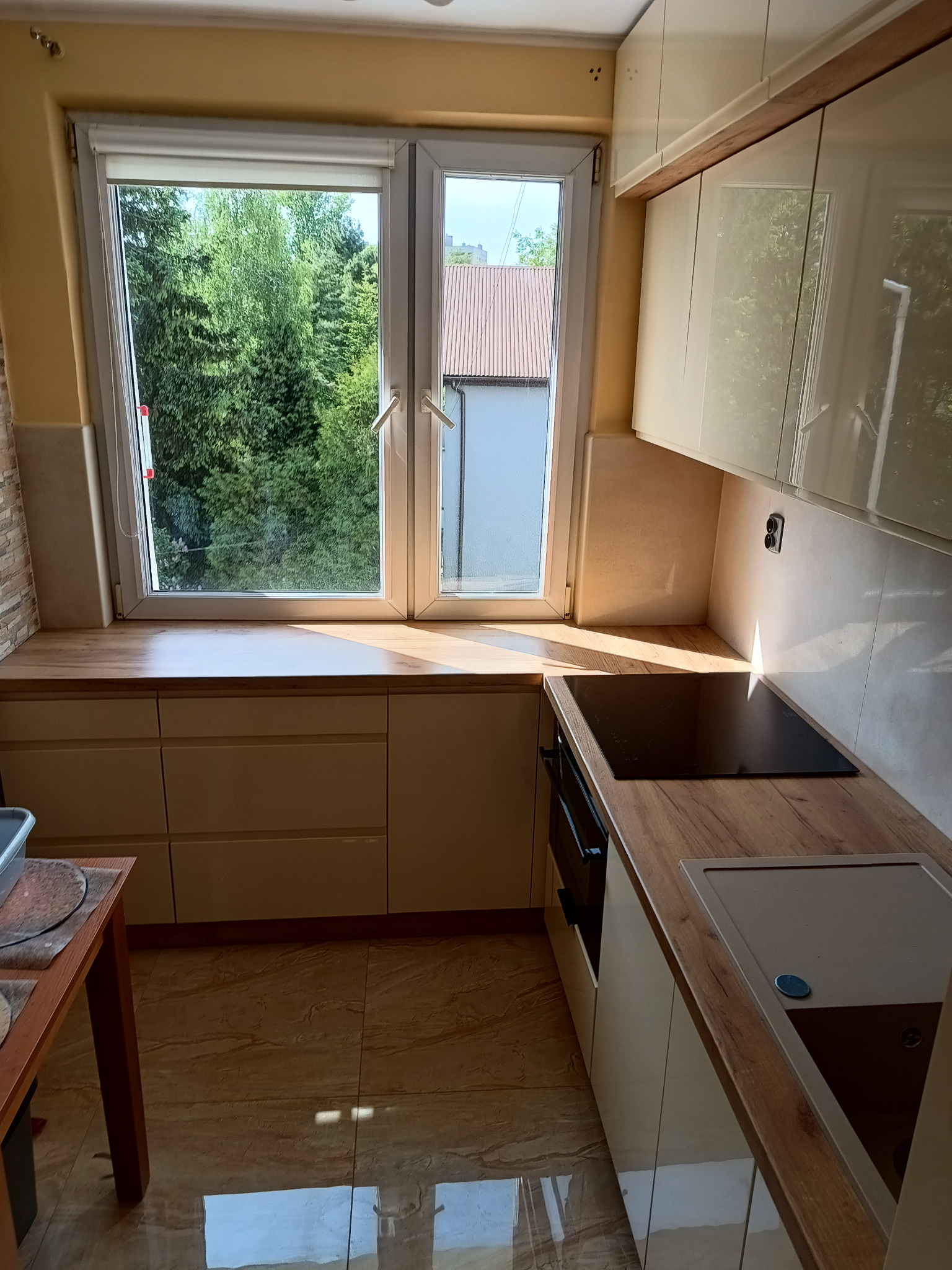 Jasna, nowoczesna kuchnia z drewnianymi blatami, widok na zieleń za oknem, płyta indukcyjna i zintegrowany z blatem zlew.