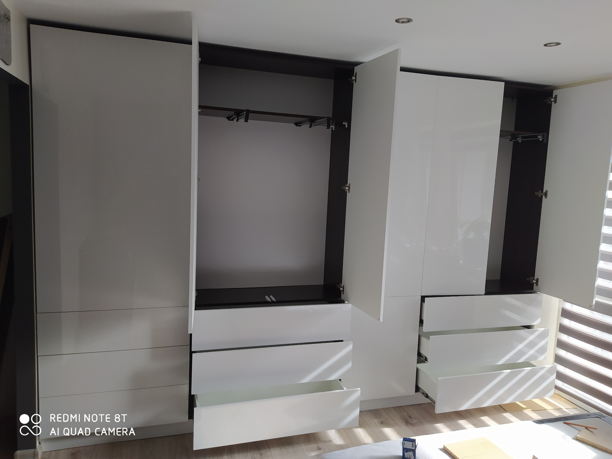 Biała, lakierowana szafa z otwartymi drzwiami i szufladami, widoczne drążki na ubrania i mechanizmy szuflad, w tle rolety okienne.