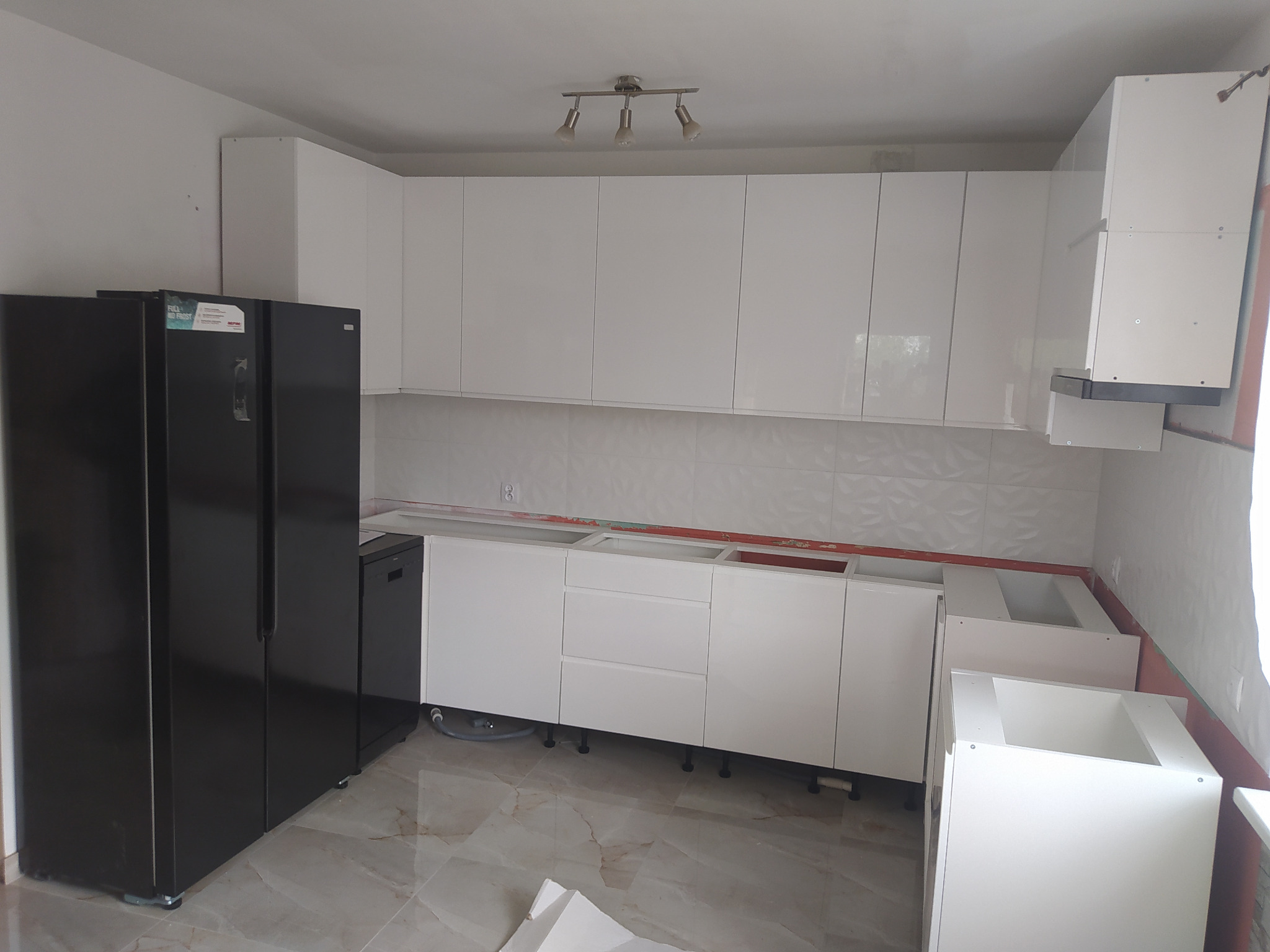 Wnętrze kuchni w trakcie montażu, widoczne białe szafki bez blatów, czarna lodówka side-by-side i fragment marmurowej podłogi.
