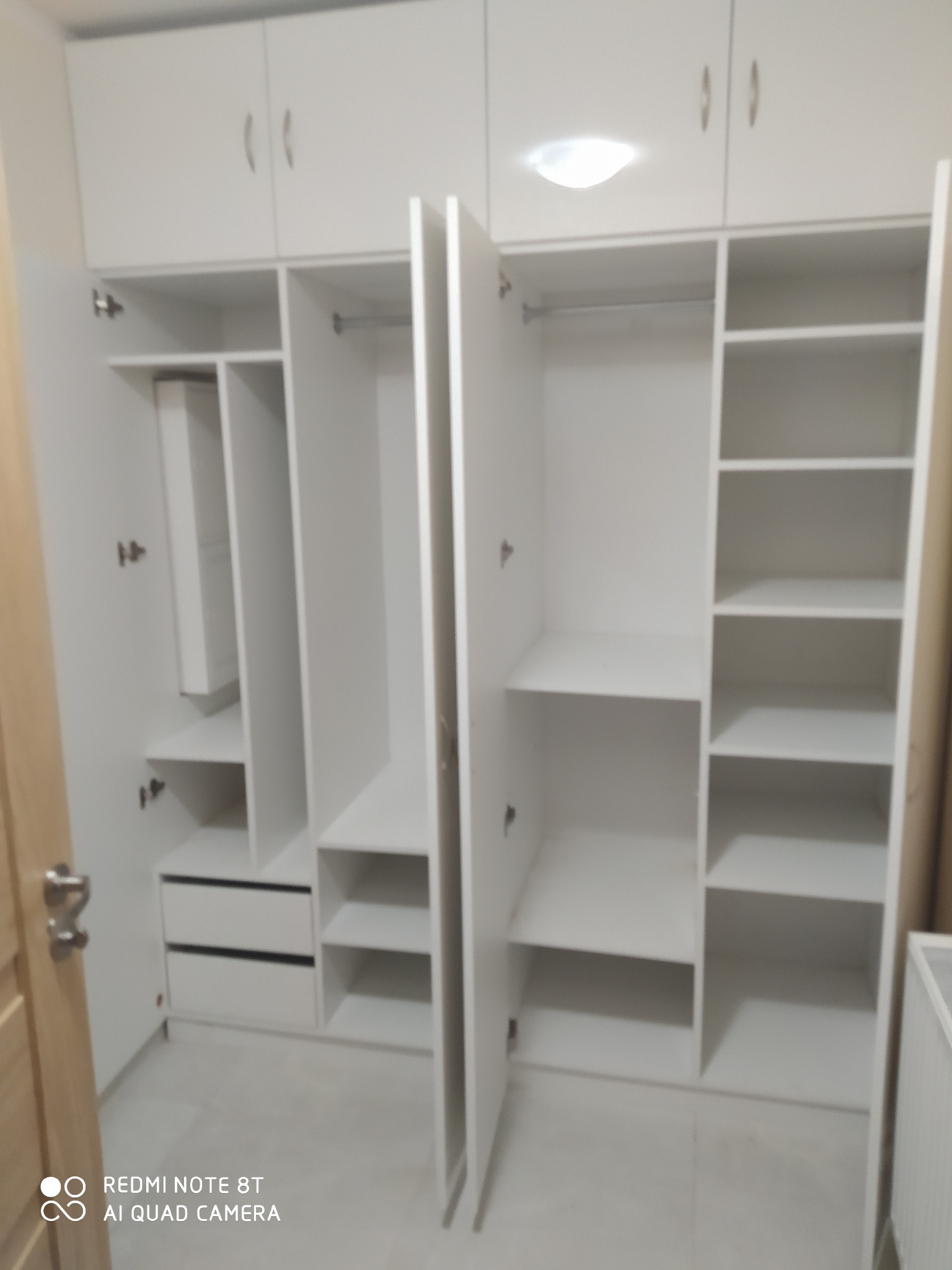 Biała szafa wnękowa z otwartymi drzwiami, widoczne półki, drążki na ubrania i dwie szuflady w dolnej części.