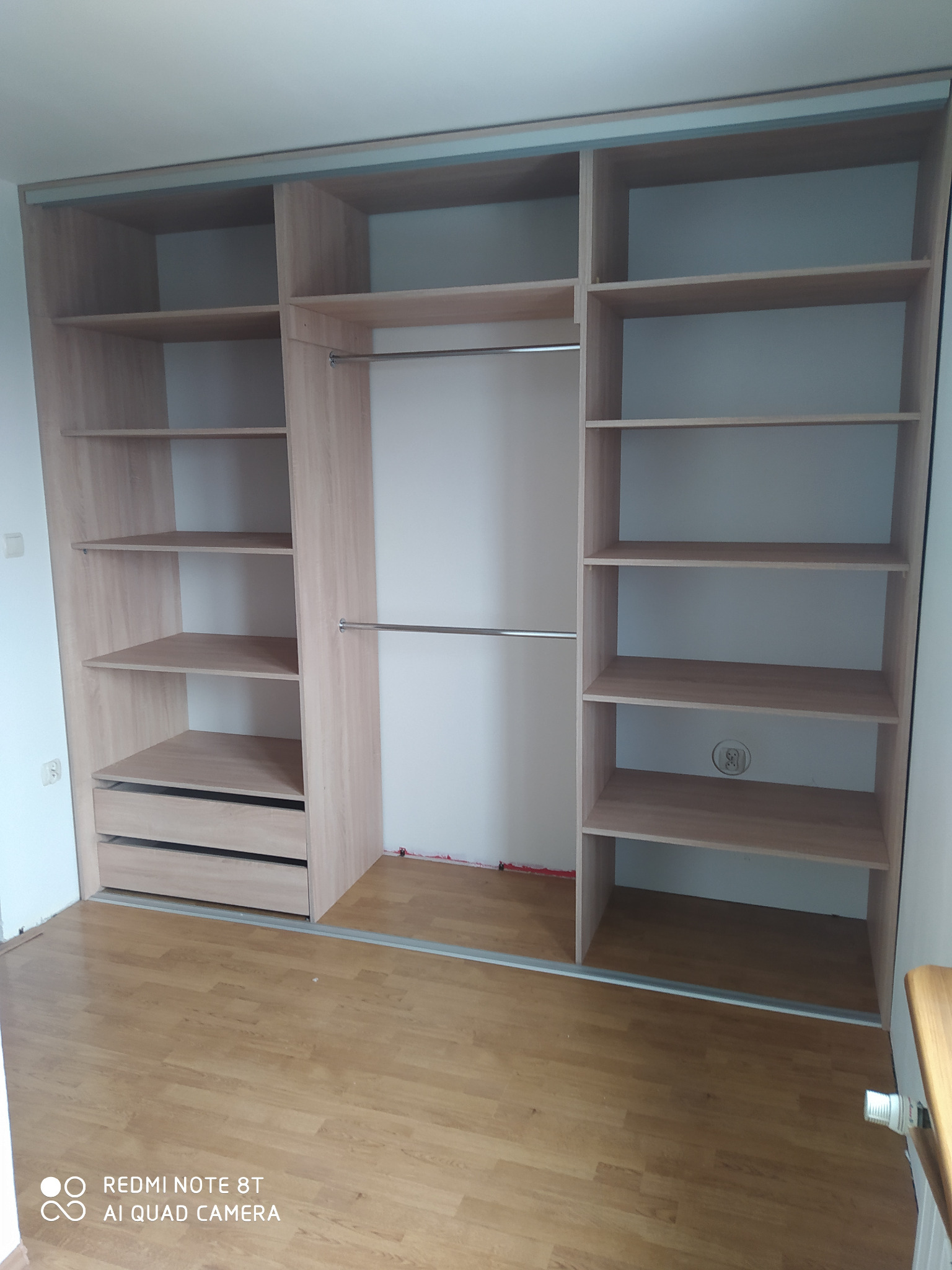 Jasna, drewnopodobna szafa wnękowa z przesuwnymi drzwiami, widoczne półki, szuflady i drążki na ubrania, zamontowana w pokoju z panelami podłogowymi.