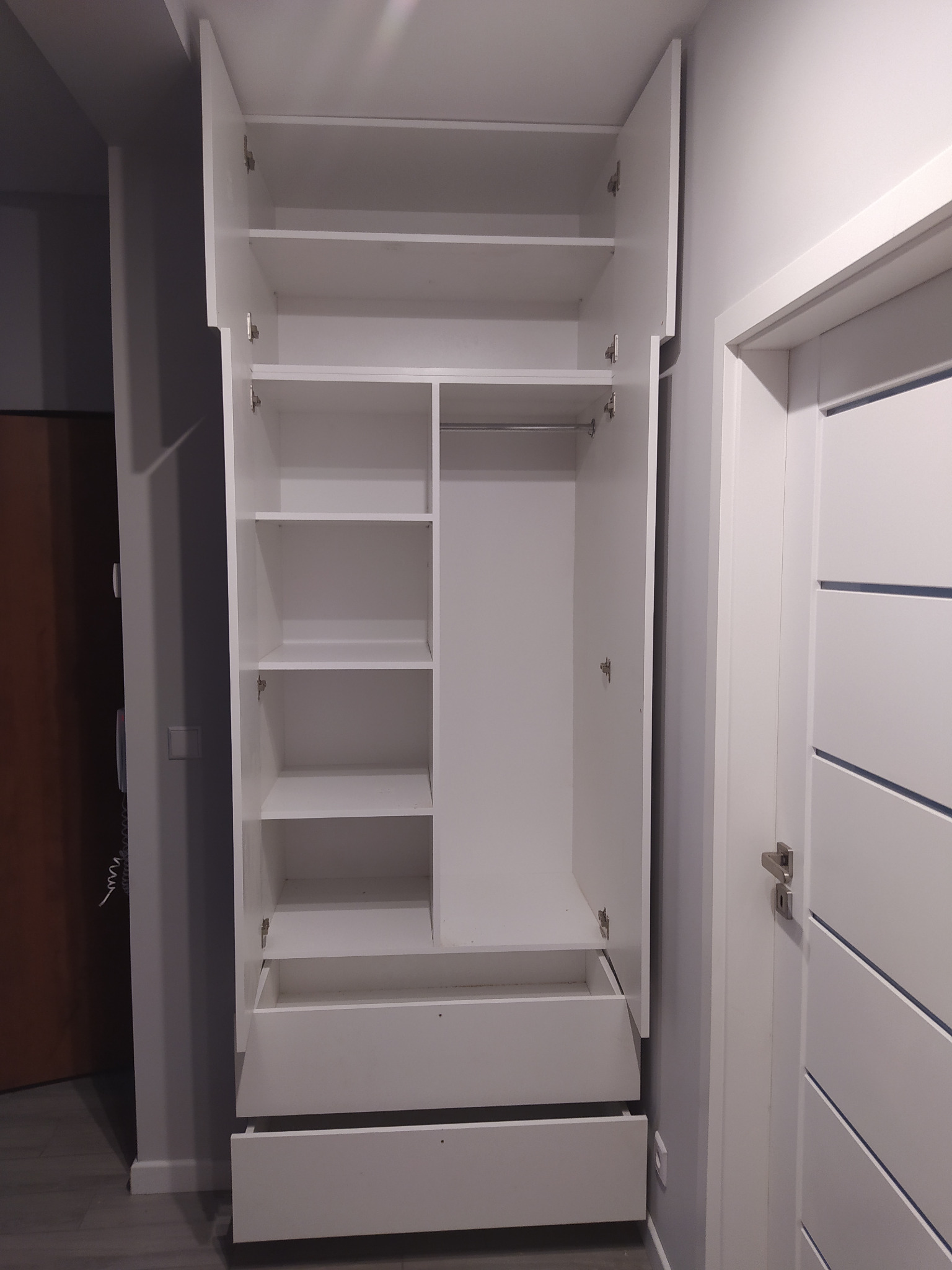 Biała szafa z otwartymi drzwiami i wysuniętymi szufladami, widoczne półki i drążek na ubrania, minimalistyczny design, szare ściany w tle.