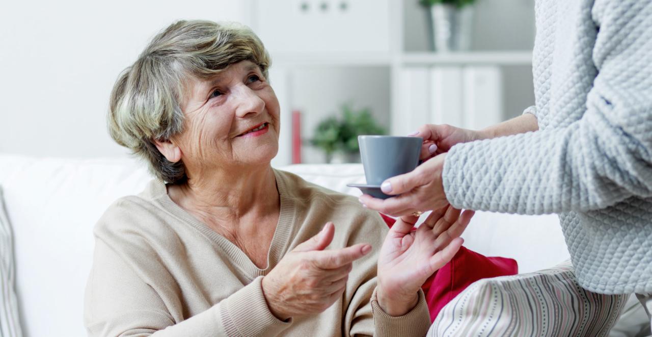 Uśmiechnięta starsza kobieta otrzymuje filiżankę herbaty od opiekuna w szarym swetrze o strukturze plastra miodu.