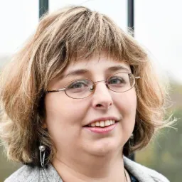 Portret uśmiechniętej kobiety w okularach z metalowymi oprawkami, krótkie blond włosy z grzywką, srebrne kolczyki z łańcuszkami, szary kardigan