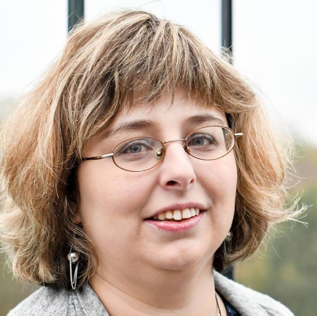 Portret uśmiechniętej kobiety w okularach z metalowymi oprawkami, krótkie blond włosy z grzywką, srebrne kolczyki z łańcuszkami, szary kardigan