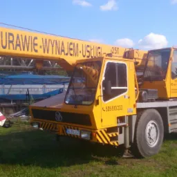 Żółty dźwig samochodowy z napisem 'Żurawie-Wynajem-Usługi 25T' na wysięgniku, stojący na trawiastym terenie w słoneczny dzień.