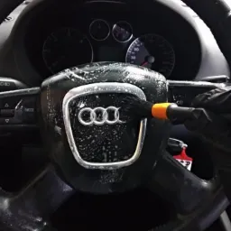 Czyszczenie kierownicy Audi z logo, pędzel z pomarańczowym uchwytem i czarna rękawica, widoczna deska rozdzielcza.