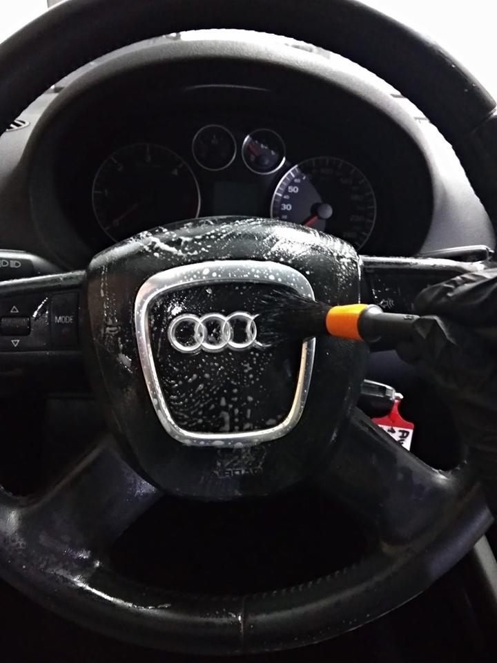 Czyszczenie kierownicy Audi z logo, pędzel z pomarańczowym uchwytem i czarna rękawica, widoczna deska rozdzielcza.