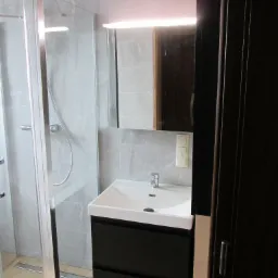 Nowoczesna łazienka z kabiną prysznicową typu walk-in, umywalką nablatową na czarnej szafce, lustrem z podświetleniem LED i szarą glazurą na ścianach.