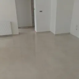 Jasne, błyszczące płytki podłogowe w nowo wykończonym pokoju z białymi ścianami i grzejnikiem; widoczne gniazdka elektryczne i fragment surowej ściany w przejściu.