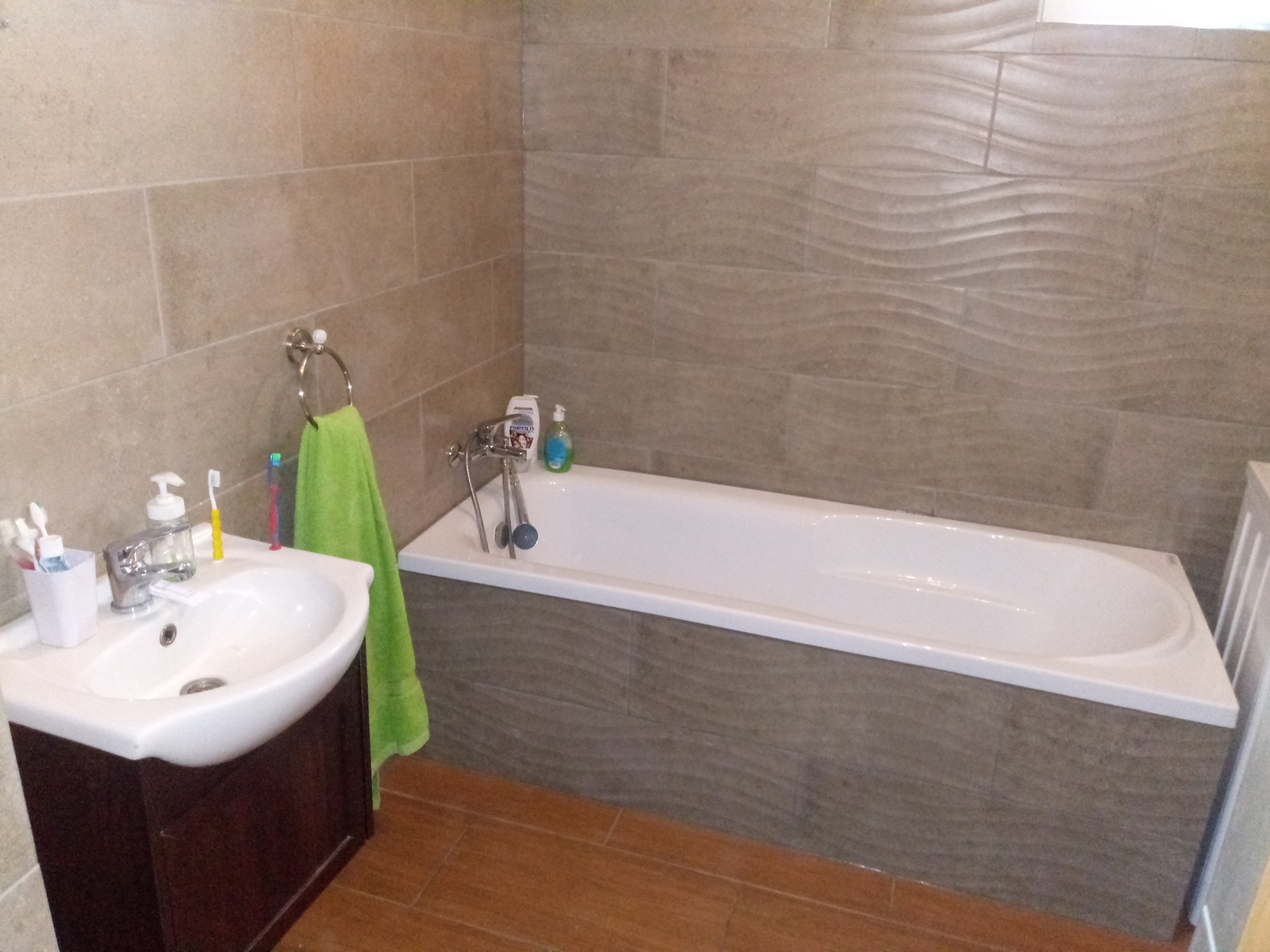Umywalka z szafką w ciemnym odcieniu drewna, biała wanna obudowana płytkami imitującymi kamień z falistą fakturą, jasnobrązowe płytki na ścianach i podłodze, zielony ręcznik na wieszaku.