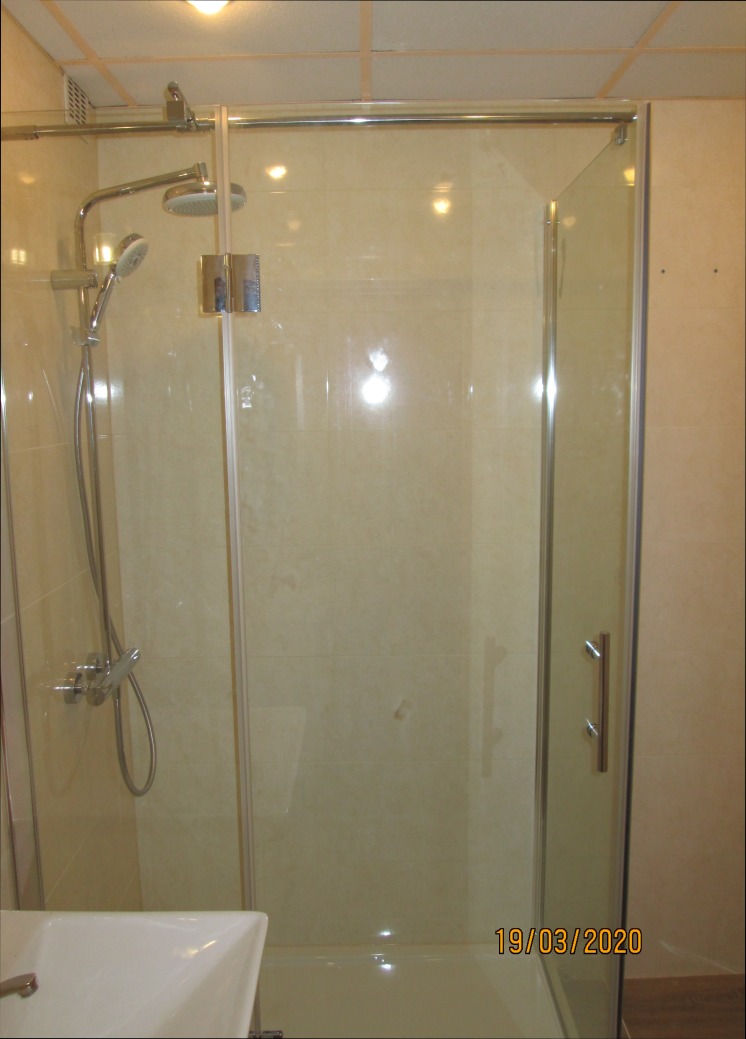 Szklana kabina prysznicowa z chromowanymi okuciami i deszczownicą, zamontowana w wykończonej łazience z beżowymi płytkami, widoczny fragment umywalki.