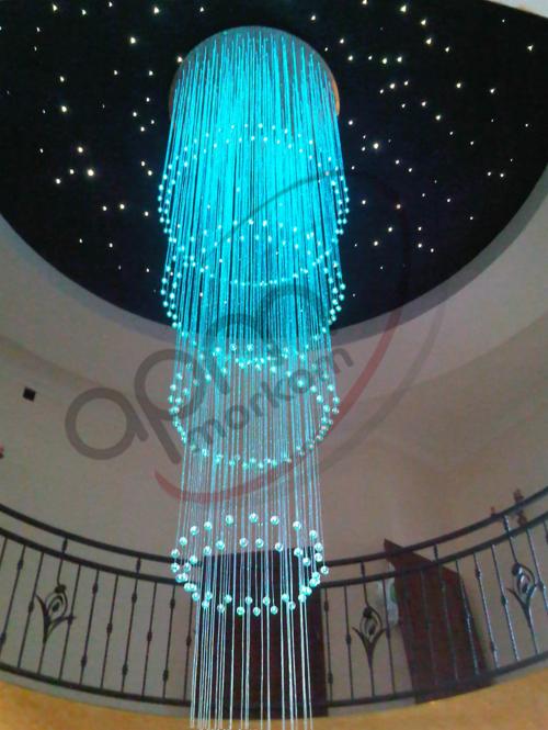 Długi, kaskadowy żyrandol z włókien optycznych w kolorze turkusowym, zwisający z sufitu imitującego nocne niebo, widoczny z dołu, z balustradą w tle.