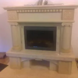 Wykończony kominek z jasnego kamienia z kolumnami w stylu greckim, widoczne detale zdobień i czarna wkładka kominkowa.