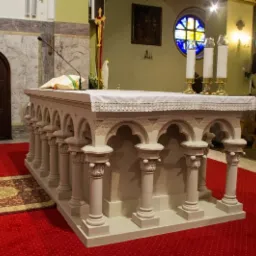 Kamienny ołtarz w kościele z kolumnami i łukami, nakryty białym obrusem, z widocznymi świecami i księgą, na czerwonym dywanie.