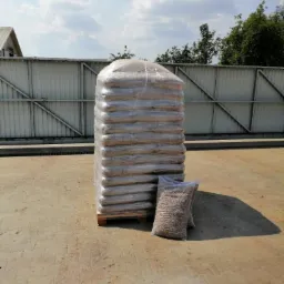 Paleta zabezpieczona folią ze stosem worków wypełnionych granulatem, ustawiona na betonowym placu w słoneczny dzień.