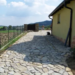 Ułożona z nieregularnych kamieni ścieżka prowadząca do domu z żółtą elewacją, widoczne elementy ogrodzenia, w tle zalesione wzgórza.