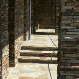 Kamienne ściany, filary i schody z nieregularnych płytek tworzące korytarz na zewnątrz budynku, oświetlone bocznym światłem słonecznym.