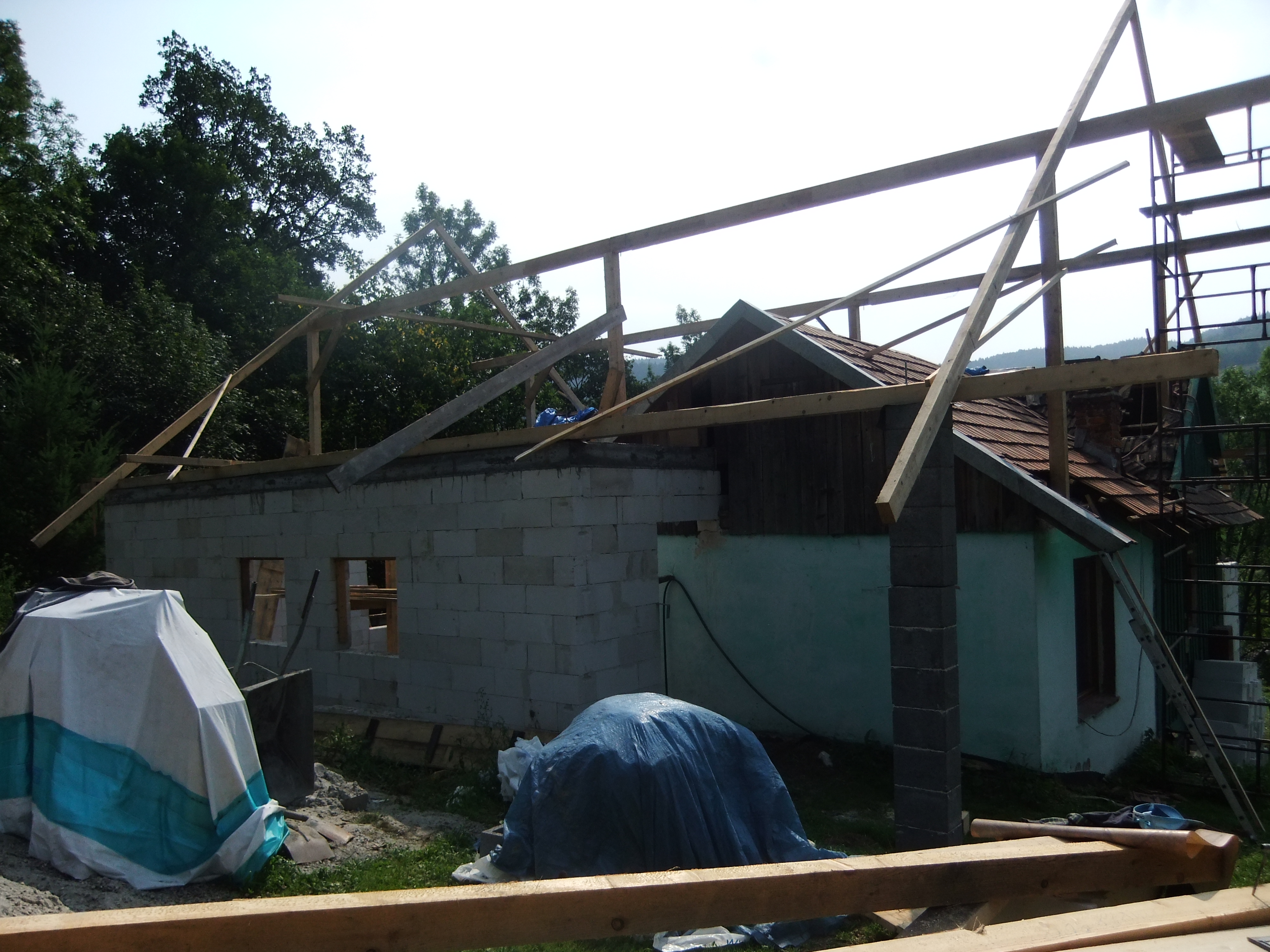 Budowa domu w trakcie: wznoszone ściany z bloczków betonowych, konstrukcja drewniana dachu w trakcie montażu, stara część budynku z czerwonym dachem widoczna w tle.