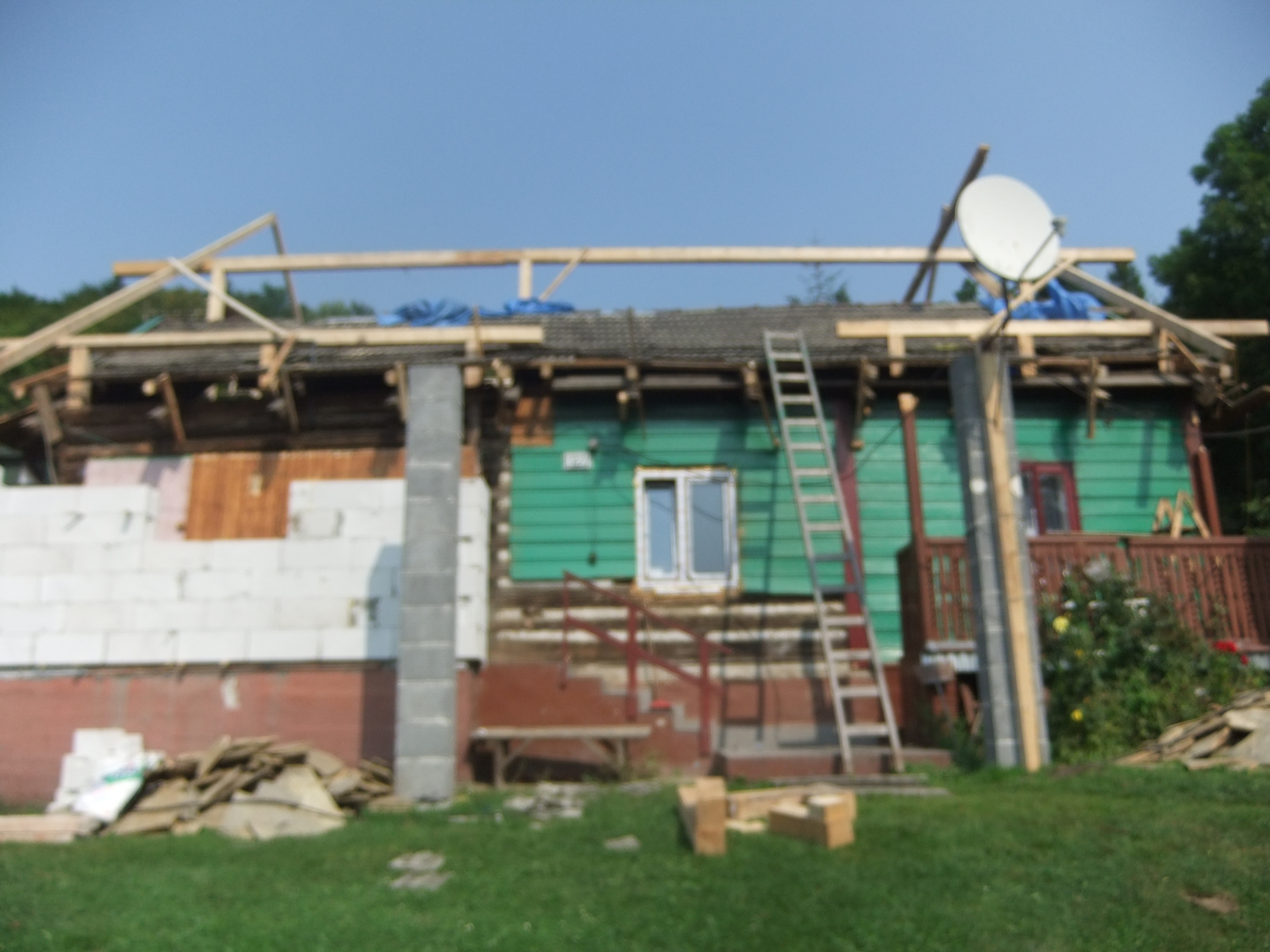 Dom w trakcie rozbudowy z widoczną konstrukcją dachu, częściowo ocieploną ścianą z białych bloczków i zieloną elewacją, z drabiną opartą o ścianę i satelitą na dachu.