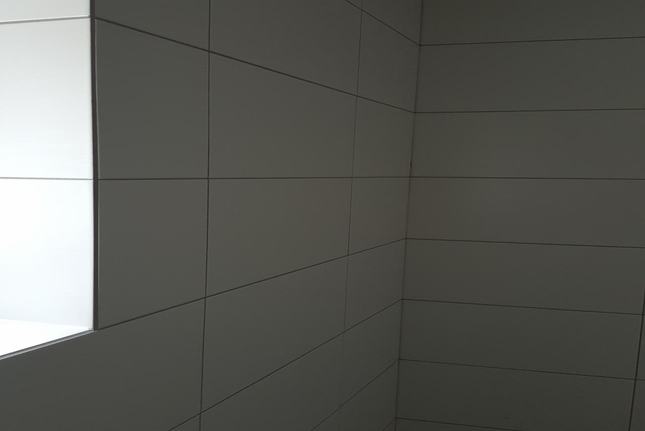 Ściana wyłożona szarymi, matowymi płytkami ceramicznymi w łazience, z widocznym fragmentem białej wnęki.
