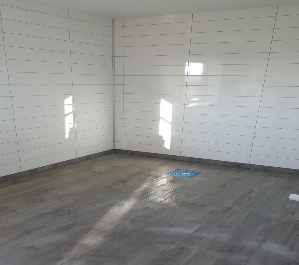 Nowoczesna łazienka po remoncie: białe, poziome płytki na ścianach z szarą fugą, podłoga imitująca drewno, niebieski odpływ liniowy w rogu.