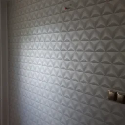 Ściana pokryta tr&oacute;jwymiarowymi panelami dekoracyjnymi w odcieniach bieli i szarości, z widocznymi elementami instalacji elektrycznej i fragmentem podłogi.