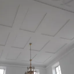 Biały sufit z kasetonami i sztukaterią, widok z dołu, w tle okna i fragment żyrandola.