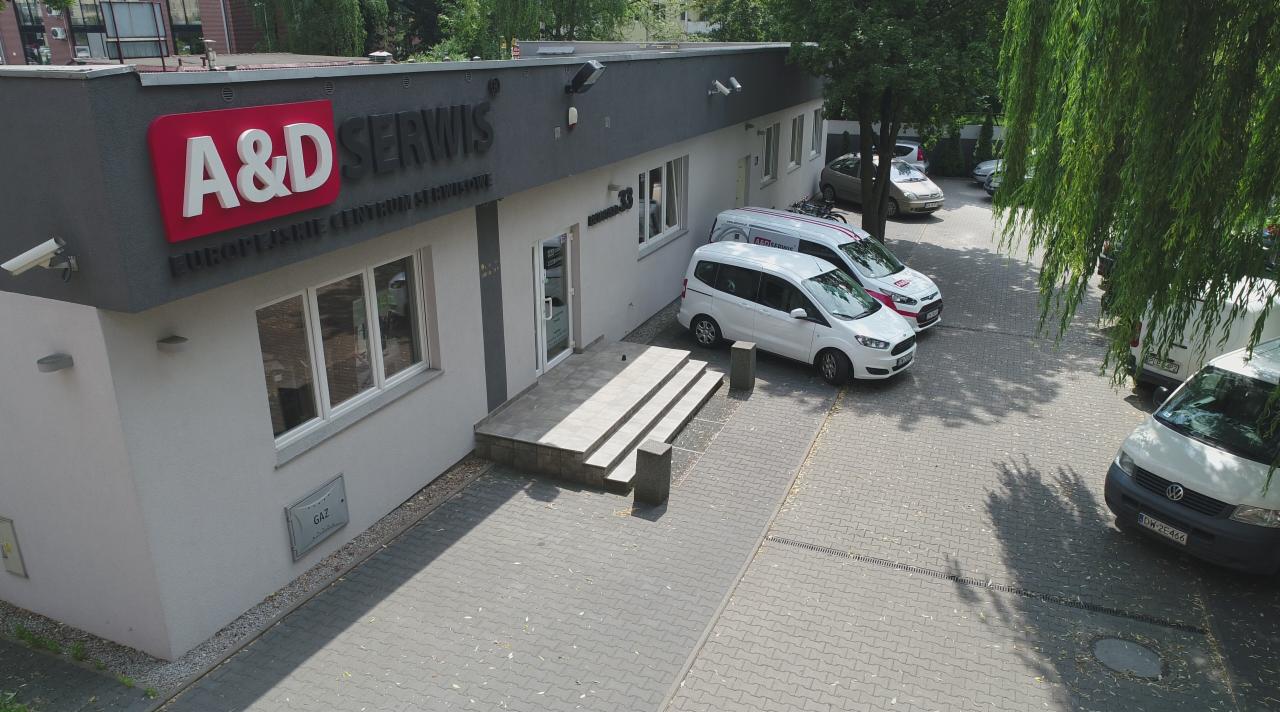 Budynek serwisu A&D z logo i zaparkowanymi białymi samochodami firmowymi na brukowanym parkingu, widok z góry.