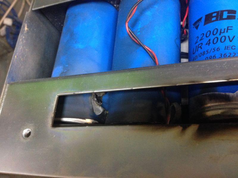 Uszkodzony kondensator elektrolityczny wewnątrz metalowej obudowy urządzenia elektronicznego, widoczne pęknięcie obudowy i ślady przepalenia.