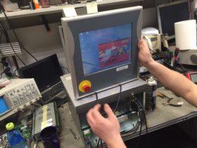 Mężczyzna podtrzymuje panel sterowania przemysłowego z widocznym ekranem i przyciskiem bezpieczeństwa, otoczony sprzętem pomiarowym i narzędziami na warsztatowym stole.