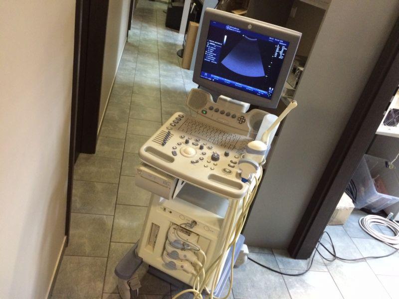 Mobilny aparat USG z widocznym ekranem i panelem sterowania, stojący na kafelkowanej podłodze w korytarzu szpitalnym.