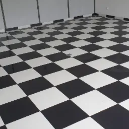 Błyszcząca, czarno-biała podłoga w szachownicę, ułożona z płytek, z widocznym odpływem liniowym w rogu pomieszczenia i szarą ścianą w tle.