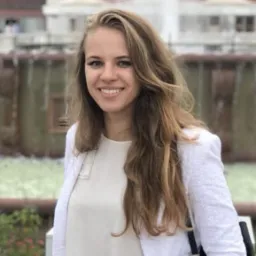 Uśmiechnięta kobieta w białej marynarce na tle fontanny i budynku z łukami.