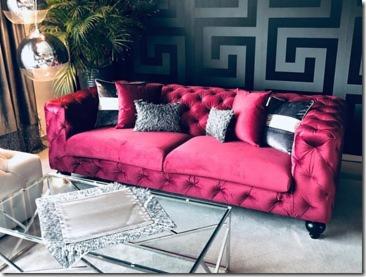 Czerwona, pikowana sofa typu chesterfield z dekoracyjnymi poduszkami w salonie z wzorzystą tapetą w odcieniach szarości i czerni. Na szklanym stoliku dekoracyjna taca. W tle widoczna roślina...