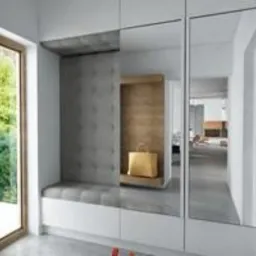 Nowoczesny przedpokój z wbudowaną szafą, lustrzanymi drzwiami i tapicerowanym siedziskiem w kolorze szarym. Na półce wnęki stoi złota torebka. Widok na jasne wnętrze mieszkania.