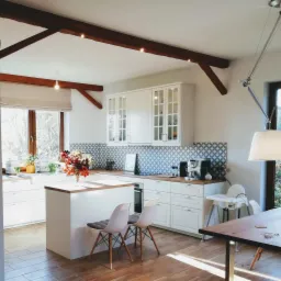 Jasna kuchnia w stylu rustykalnym z białymi meblami, drewnianymi belkami stropowymi i wzorzystą mozaiką nad blatem; na wyspie kuchennej bukiet kwiatów w wazonie.